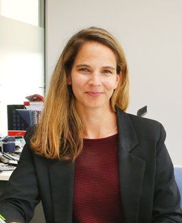 A woman is sitting at a desk wearing a black jacket and a red sweater.