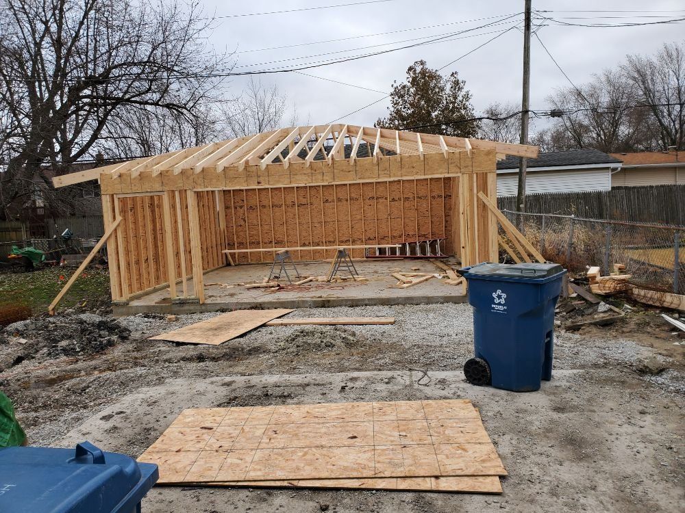 Garage under construction with wooden frame, blue trash bin, and overcast sky.