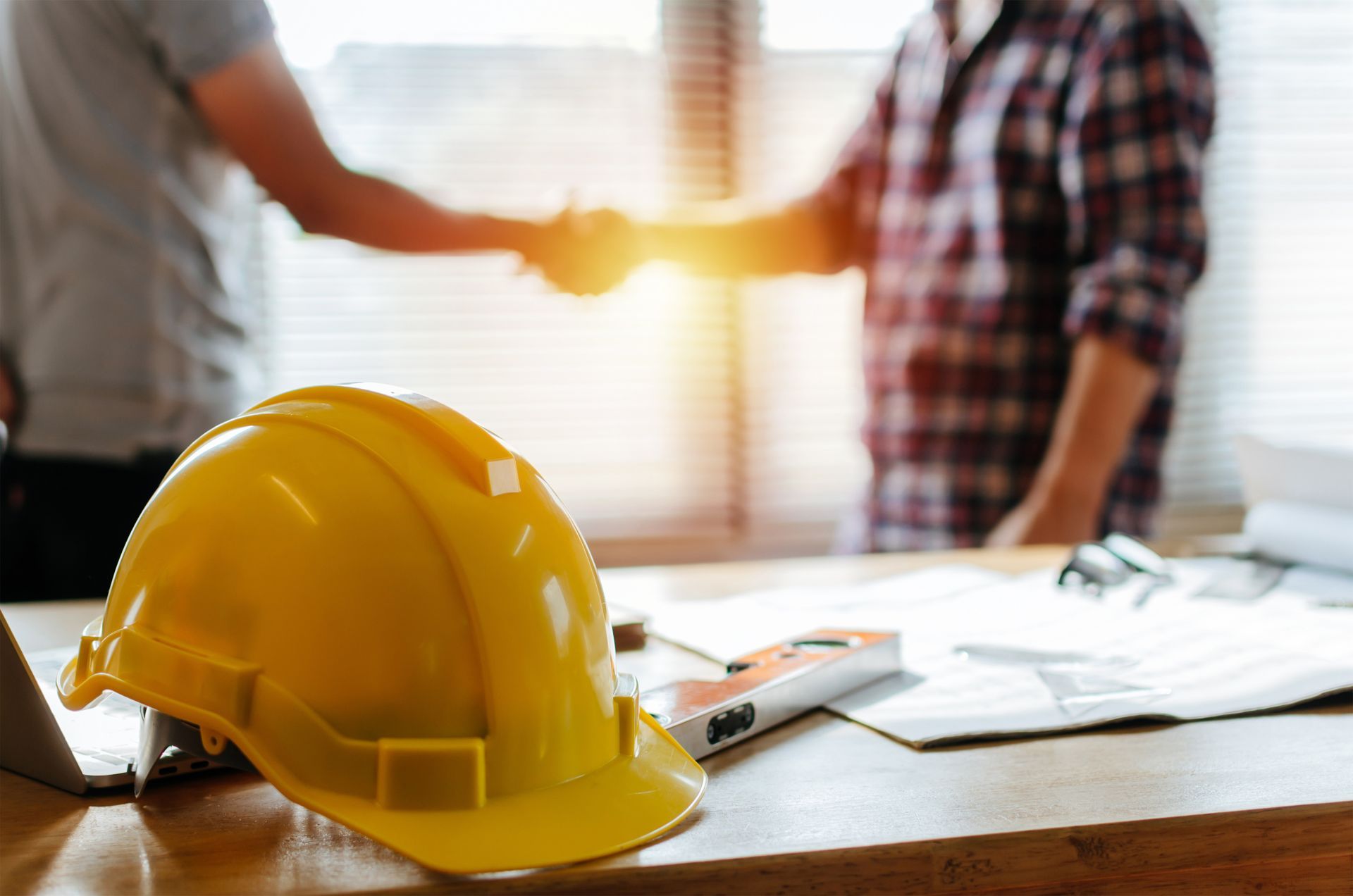 Yellow hard hat on desk; two men shaking hands in the background, possibly sealing a deal.