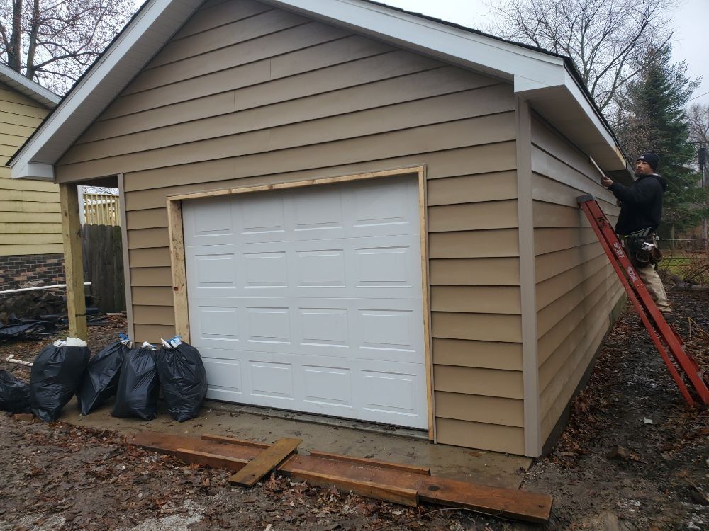 A person on a ladder is working on the siding of a tan-colored garage.