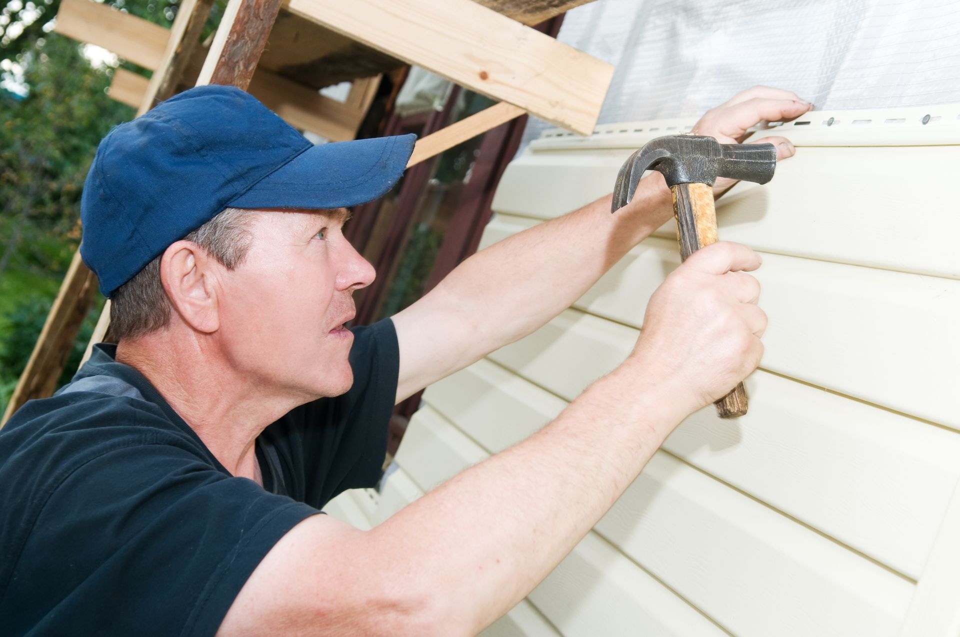 Man in blue cap hammering siding on a building's exterior.