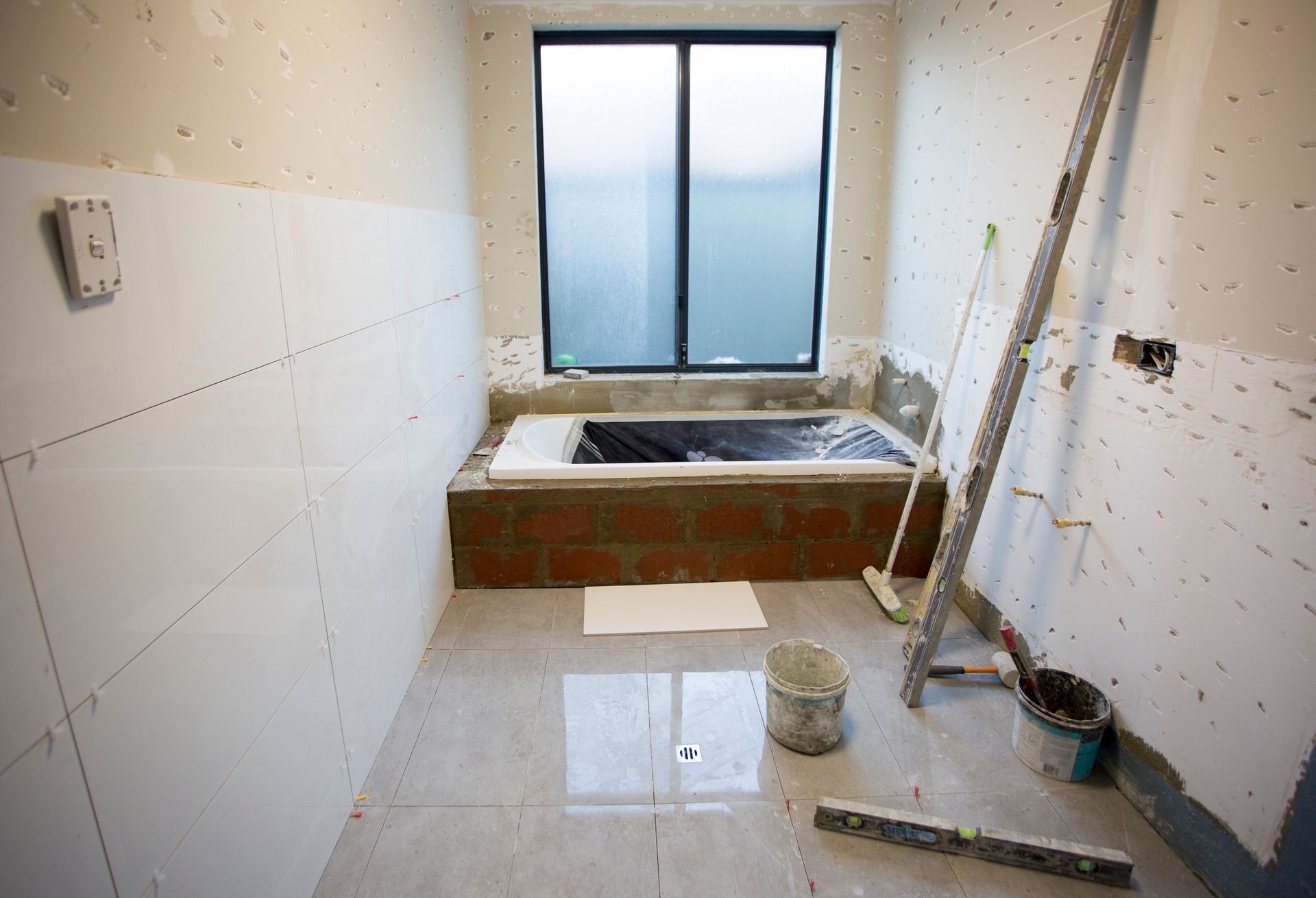 Bathroom under construction with tiled walls and floor, window, and unfinished bathtub.