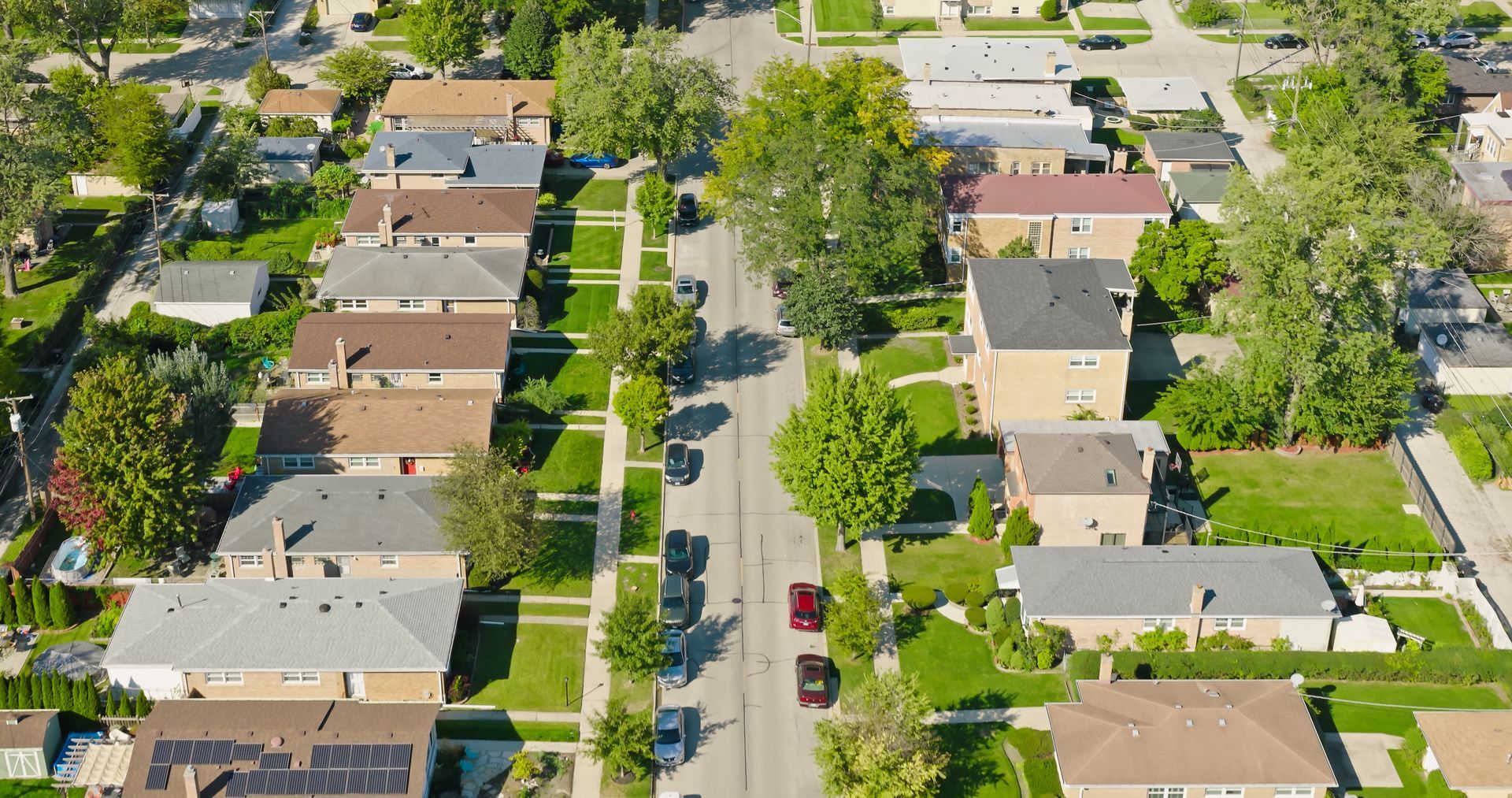 Aerial view of a suburban street lined with houses, trees, and parked cars on a sunny day.