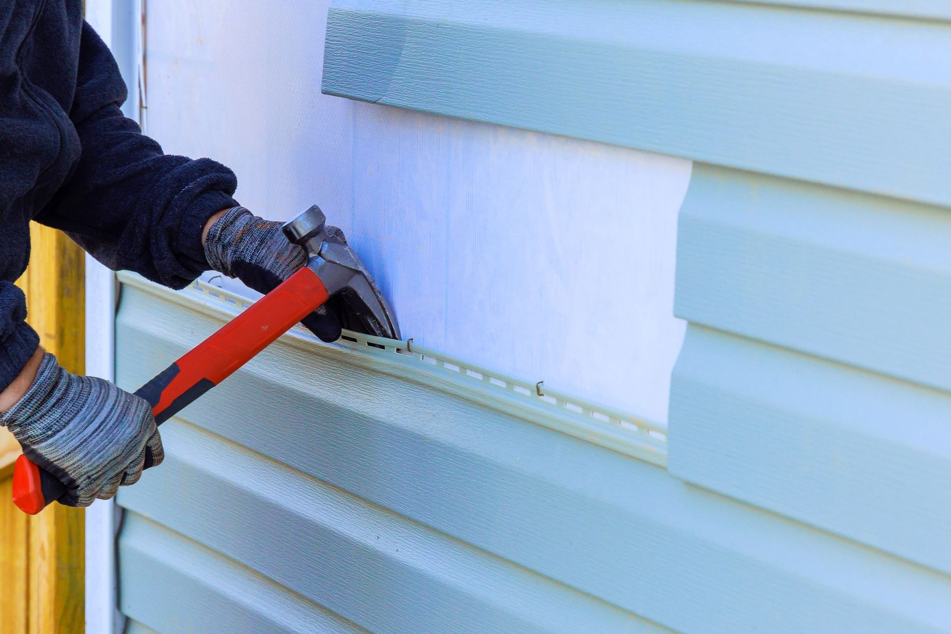 Person hammering on light blue vinyl siding with a red and black hammer.