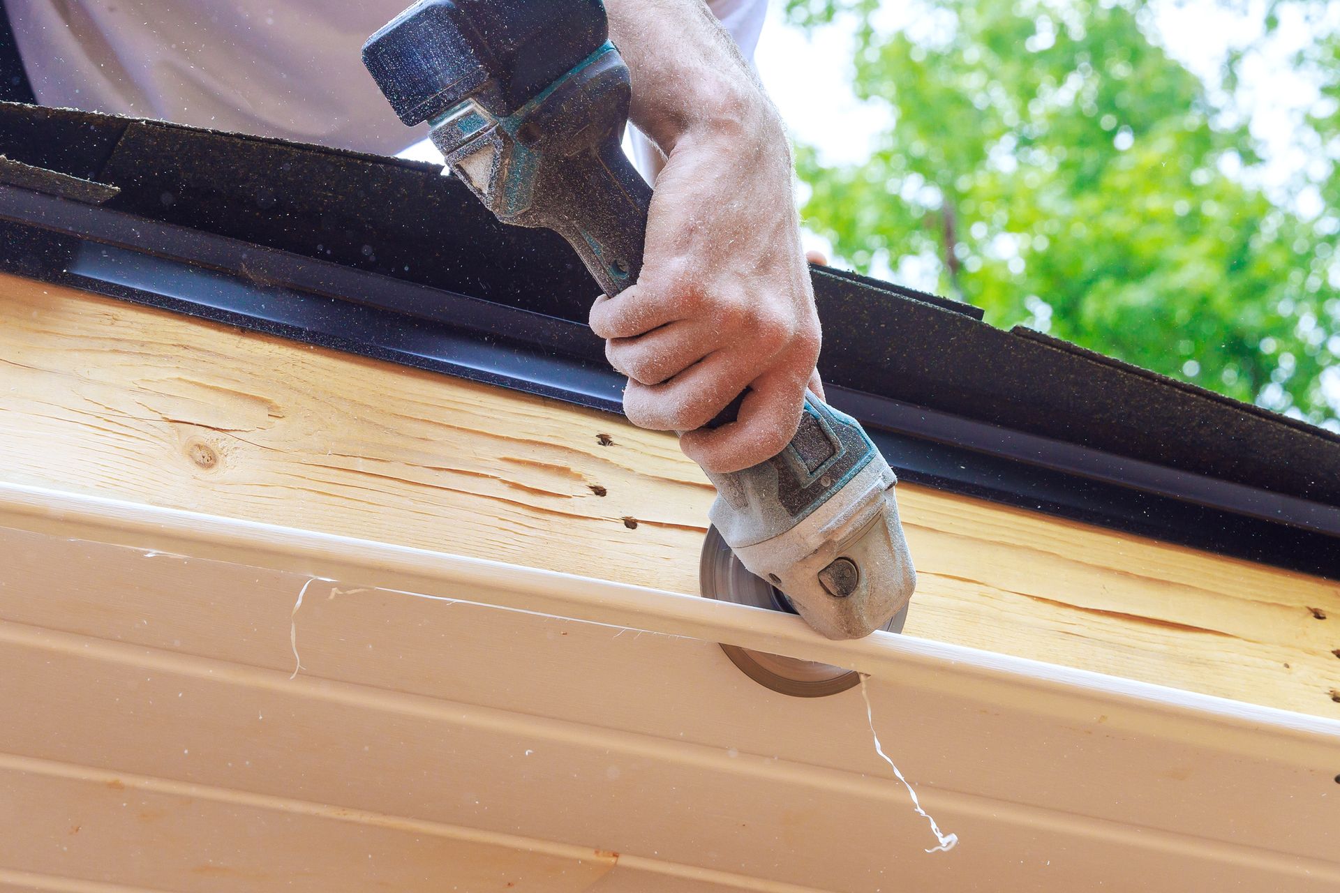 Person uses power tool to cut a white gutter. Wood beams and green foliage in background.