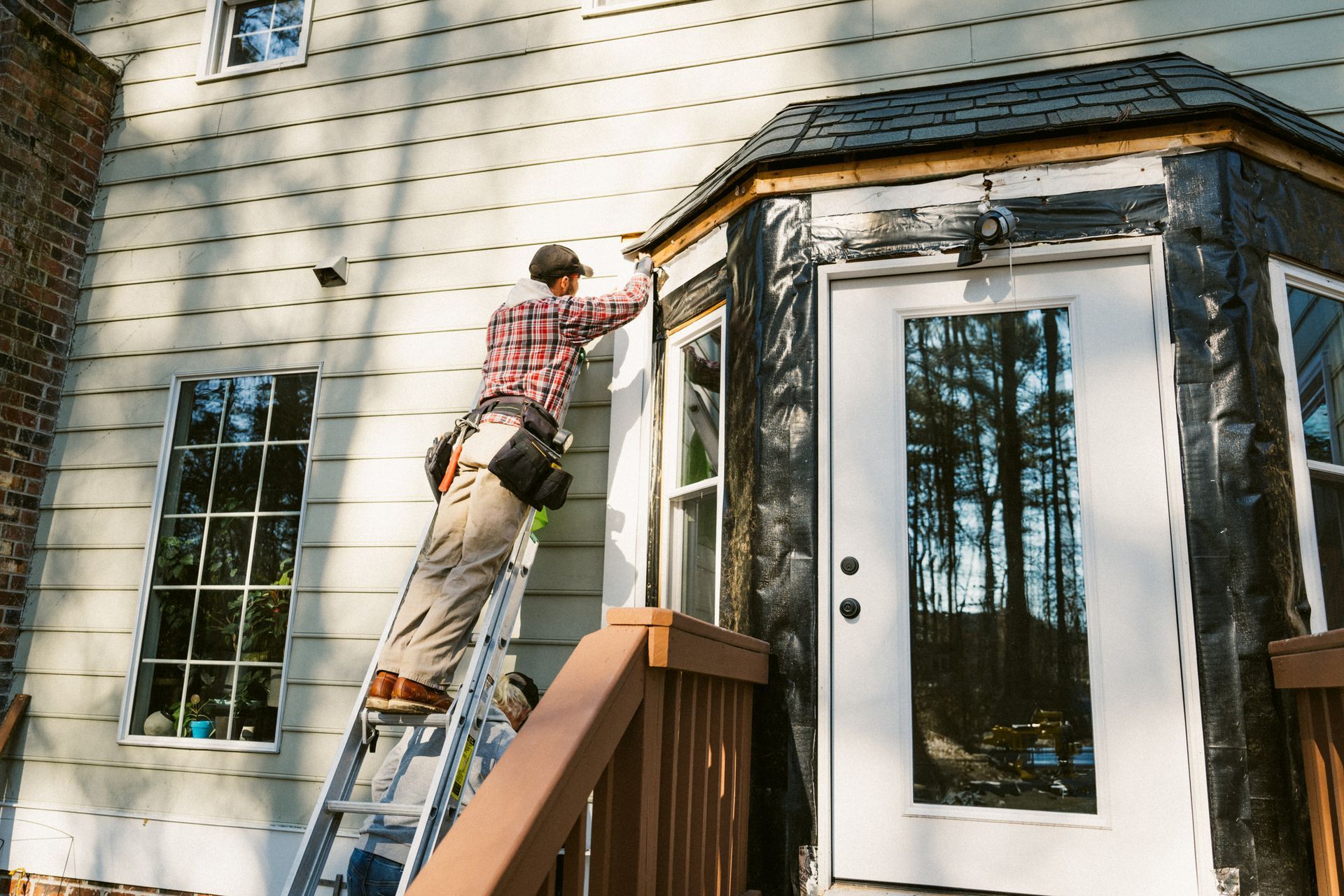A person on a ladder working on the exterior of a house, near a door and windows.