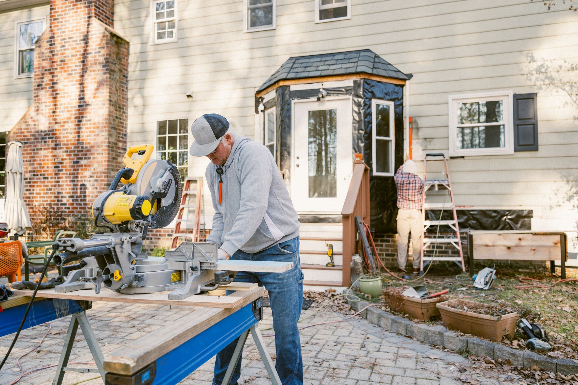 A person using a yellow and black miter saw to cut wood outdoors near a house.