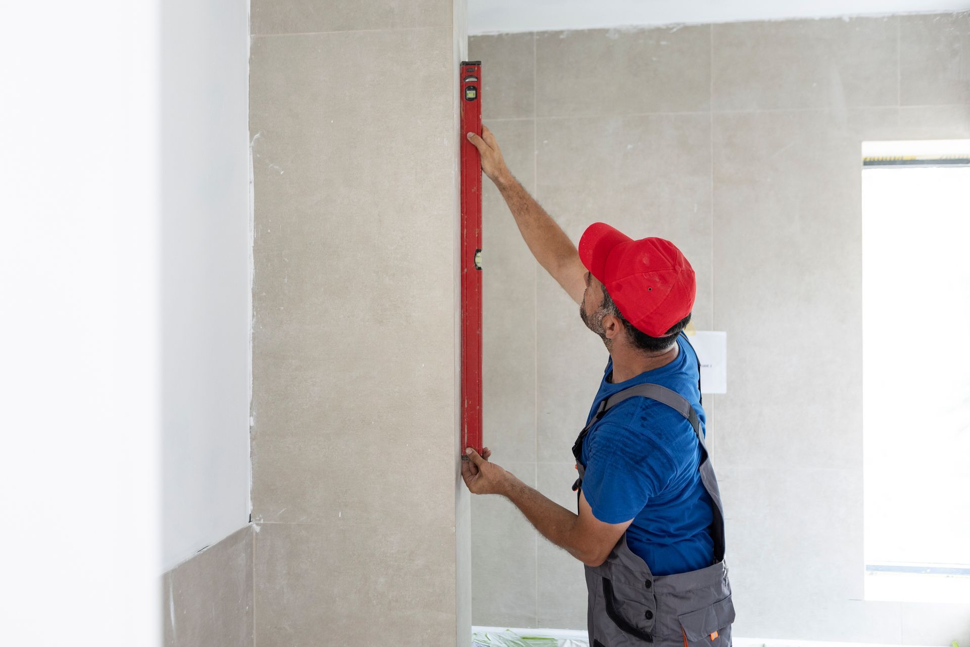 Person in overalls and red cap using a level on a wall in a room.