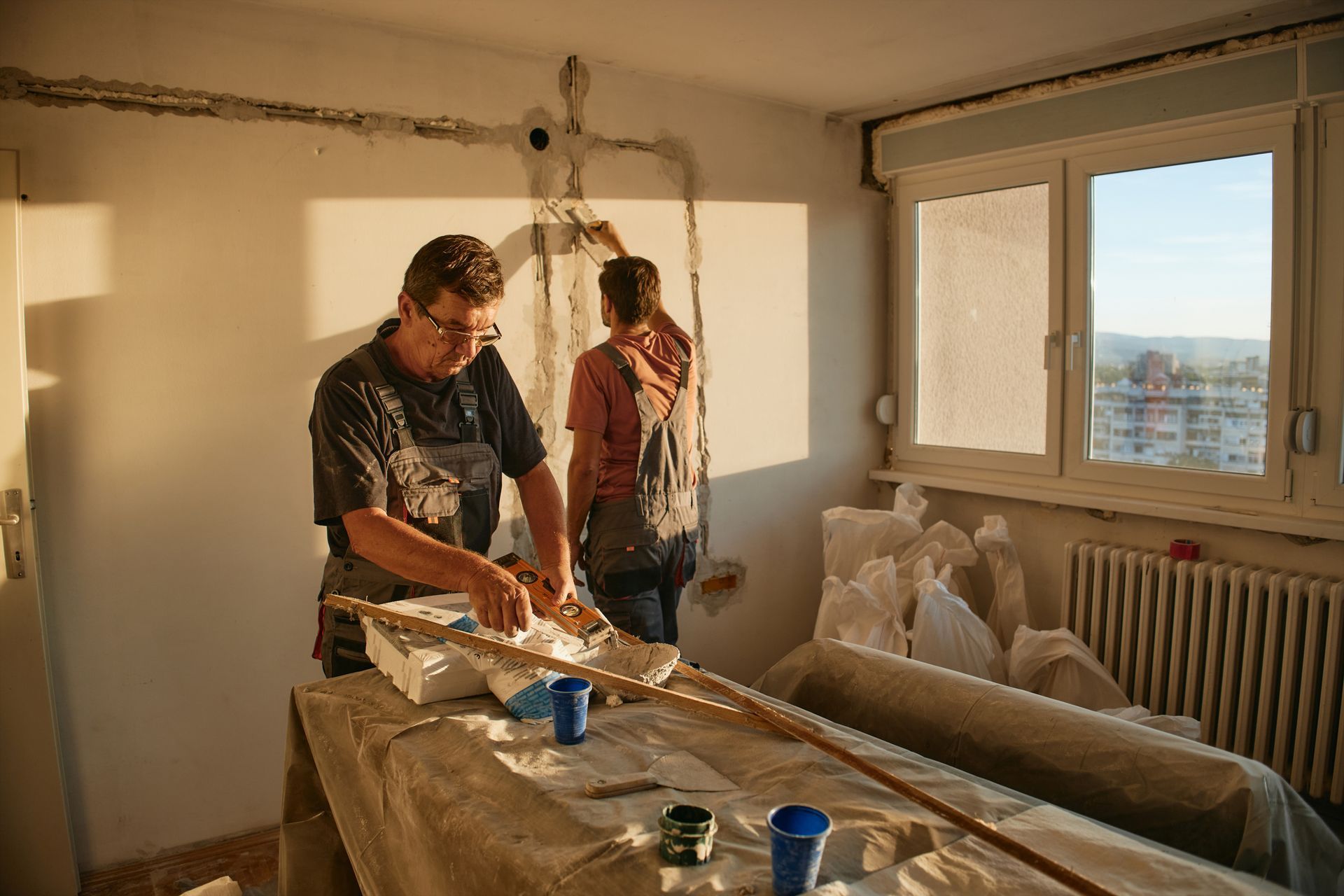 Two people renovating a room, working on a wall near a window. Sunlight streams in.