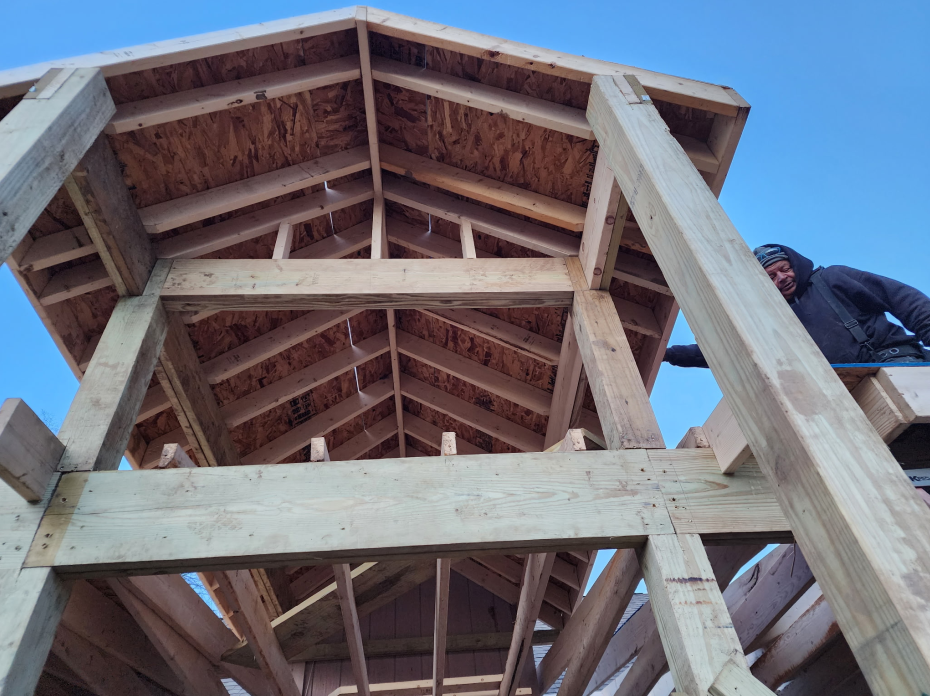 Roofer in overalls and hard hat uses a power drill on a brown metal roof under a blue sky.
