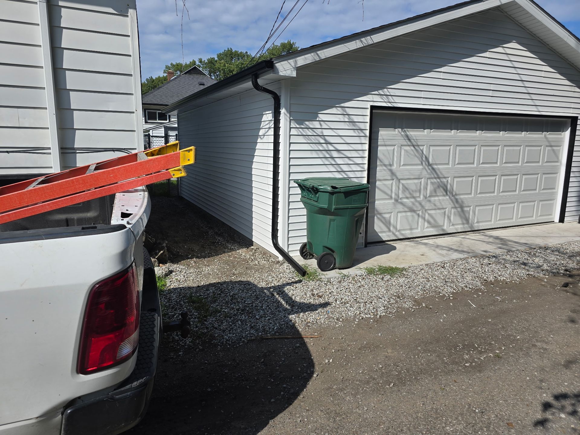 White truck with ladder next to a white garage, green trash bin on gravel.