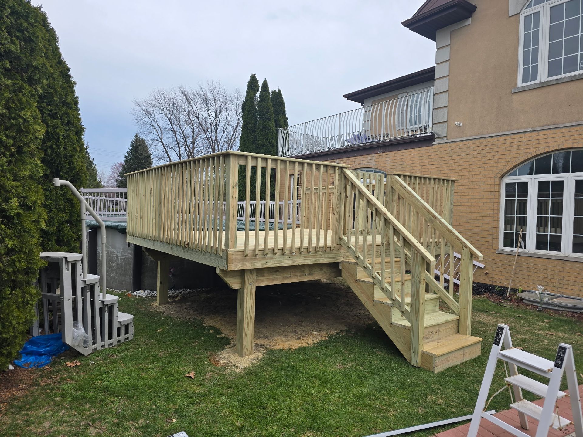 Wooden deck with stairs attached to a two-story beige house. Green grass and trees surround it.