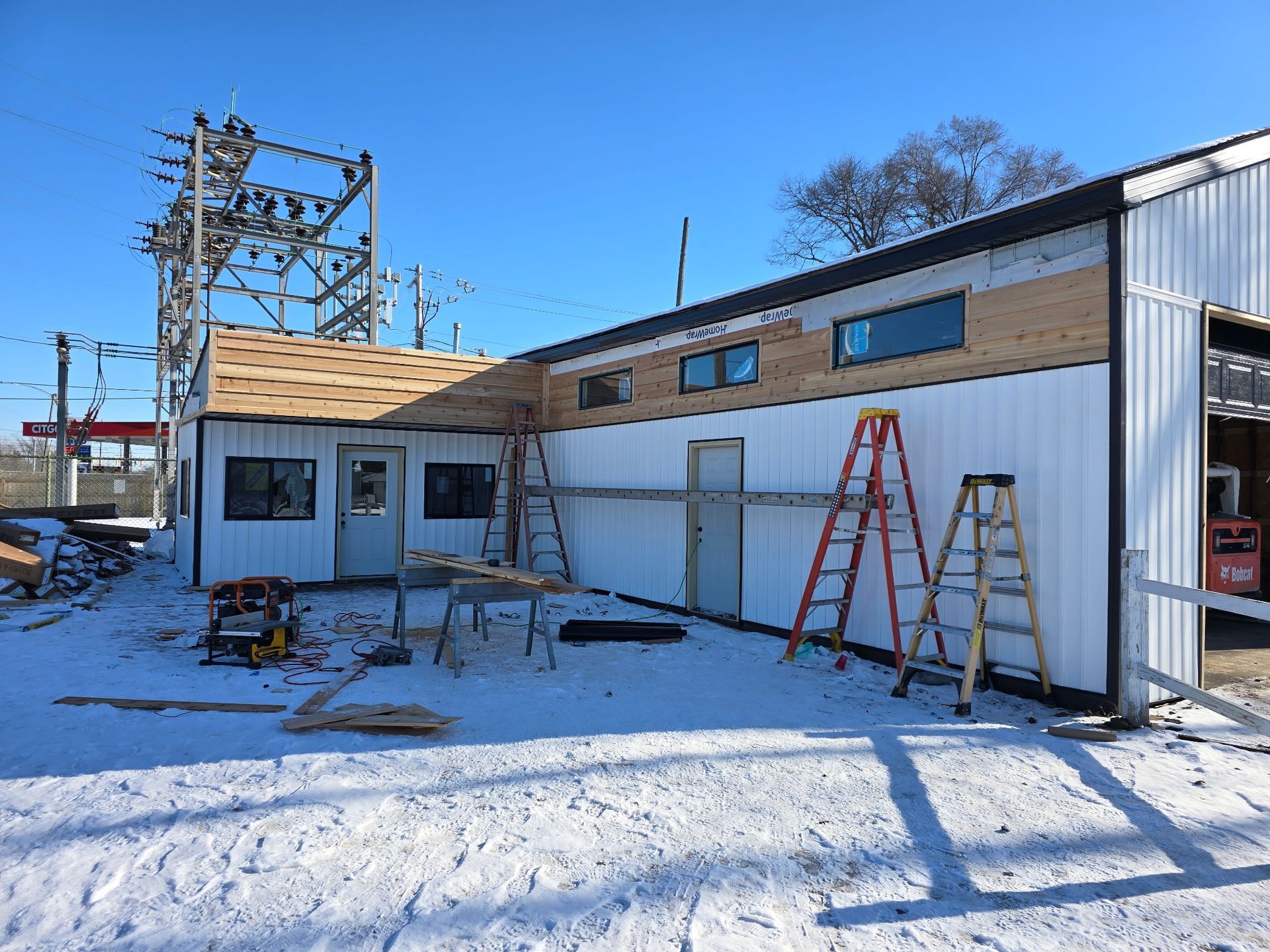 Construction site: White building with black trim, ladders, and snow-covered ground. Blue sky and power lines in the background.