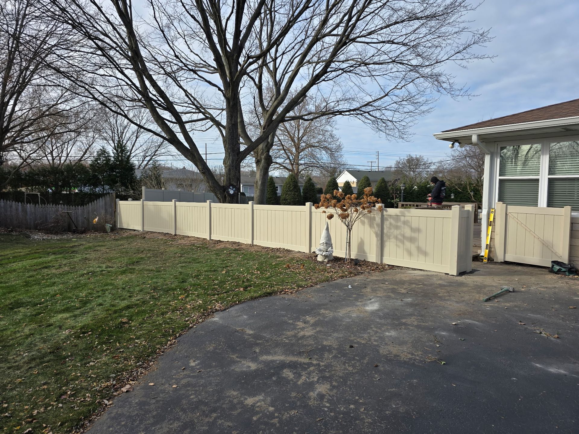 Beige fence borders a yard with a driveway. Tree in the background. Sunny day.