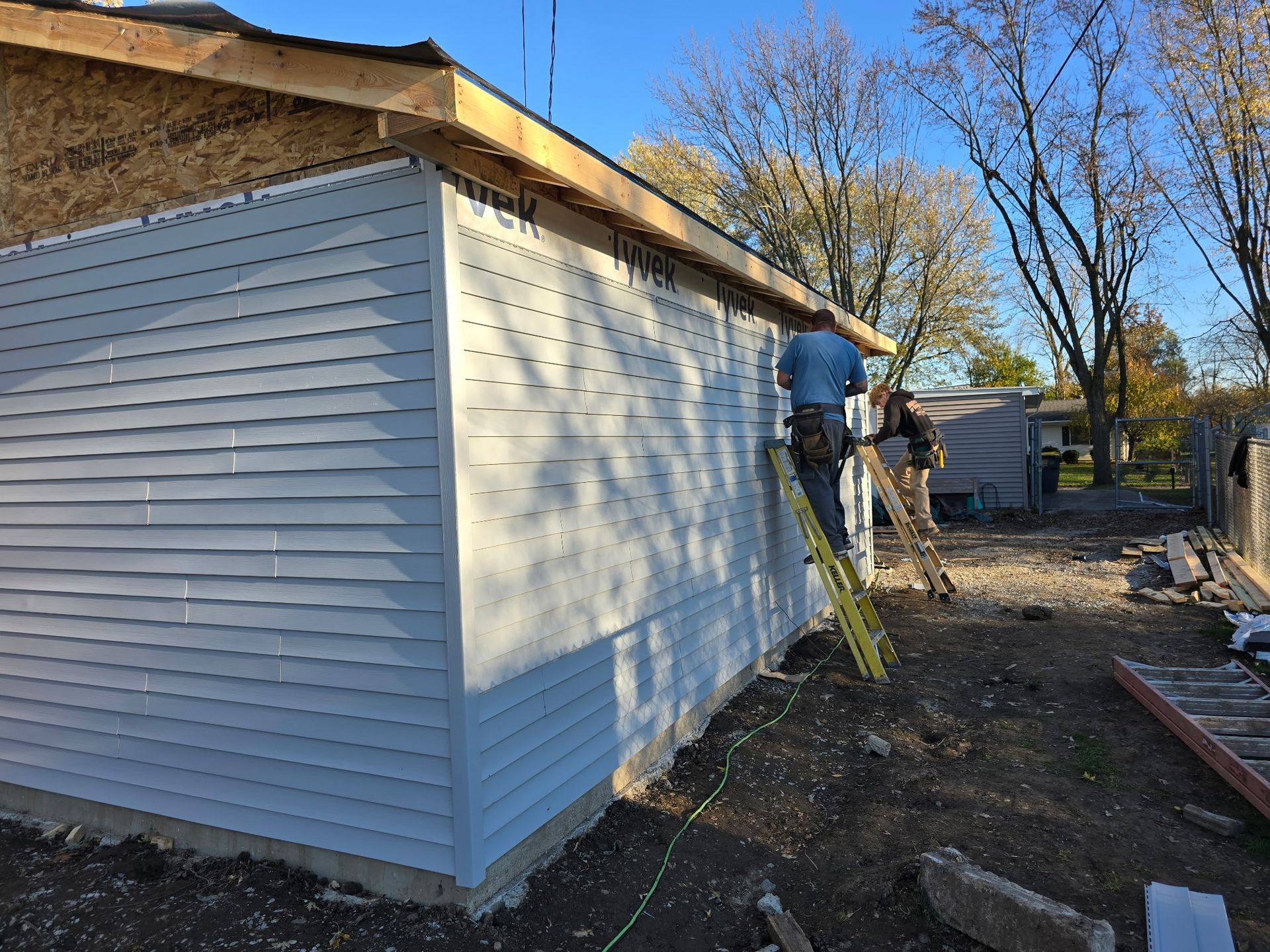 Workers install light blue siding on a building's exterior, with a ladder and a partly wooded background.