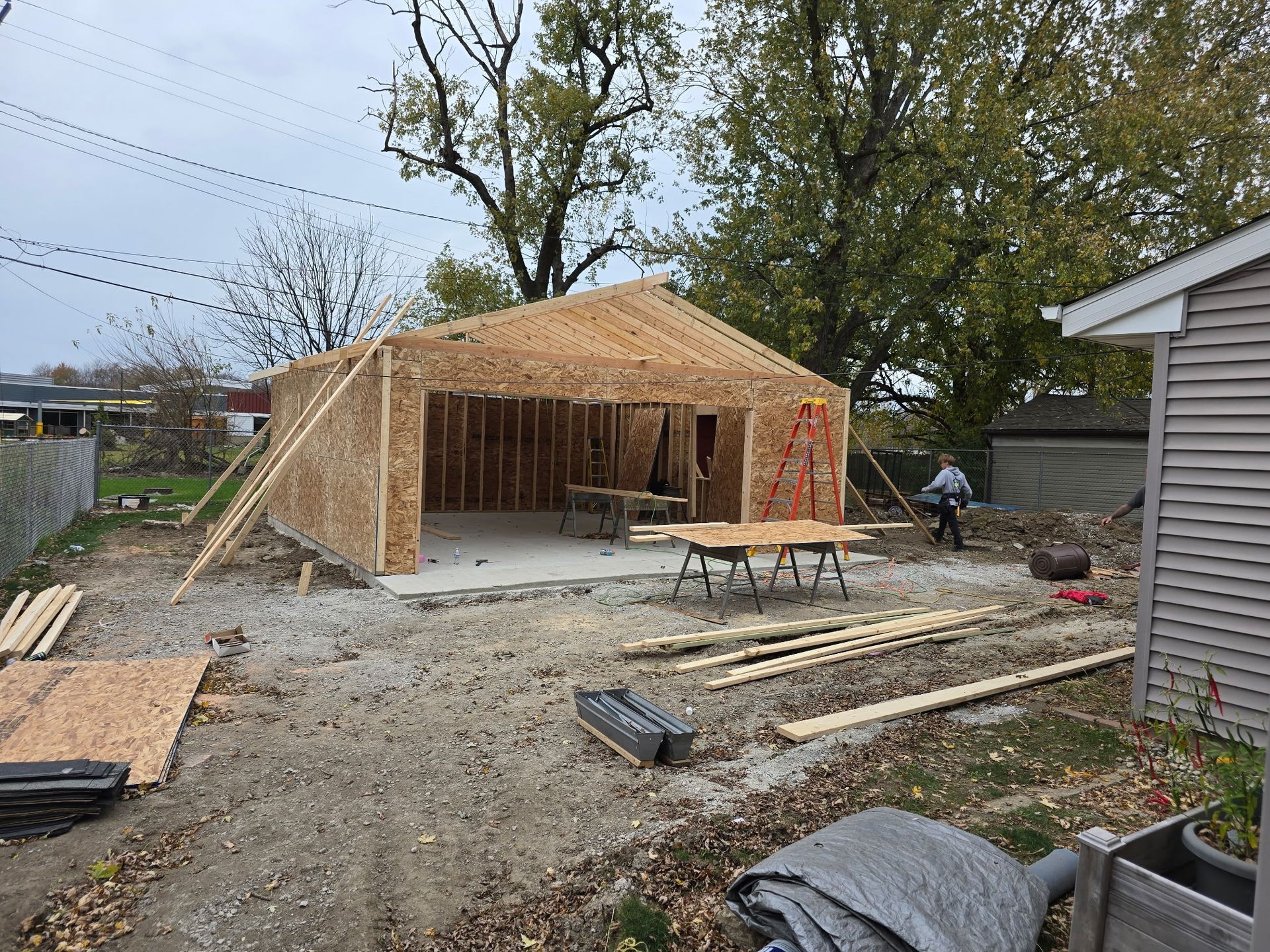 Garage under construction, wood frame, concrete floor. Person working outside next to a house. Cloudy day.
