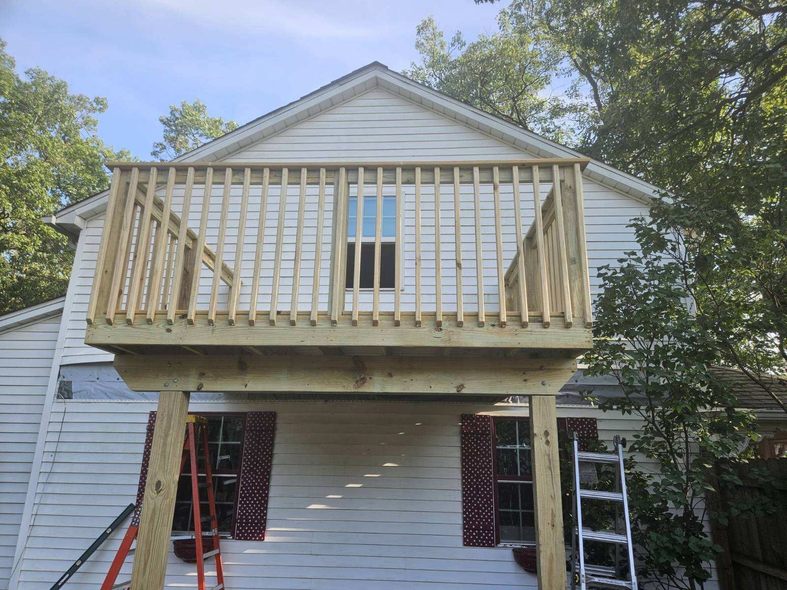 Wooden deck built on the side of a white house, with a window and railing.  A ladder is propped up to the deck.