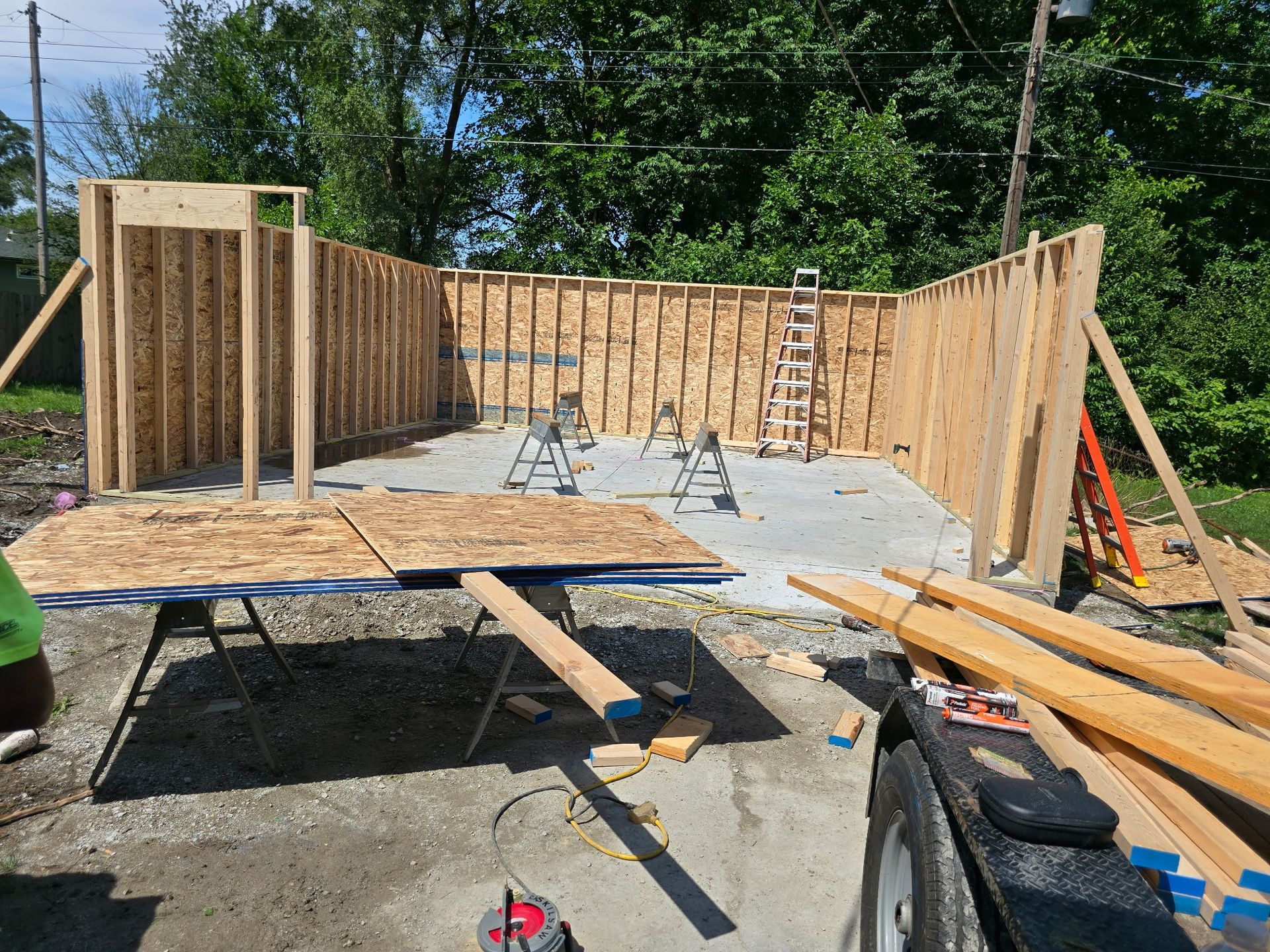 Wooden framework of a building under construction, outdoors on a concrete base, with tools and lumber visible.