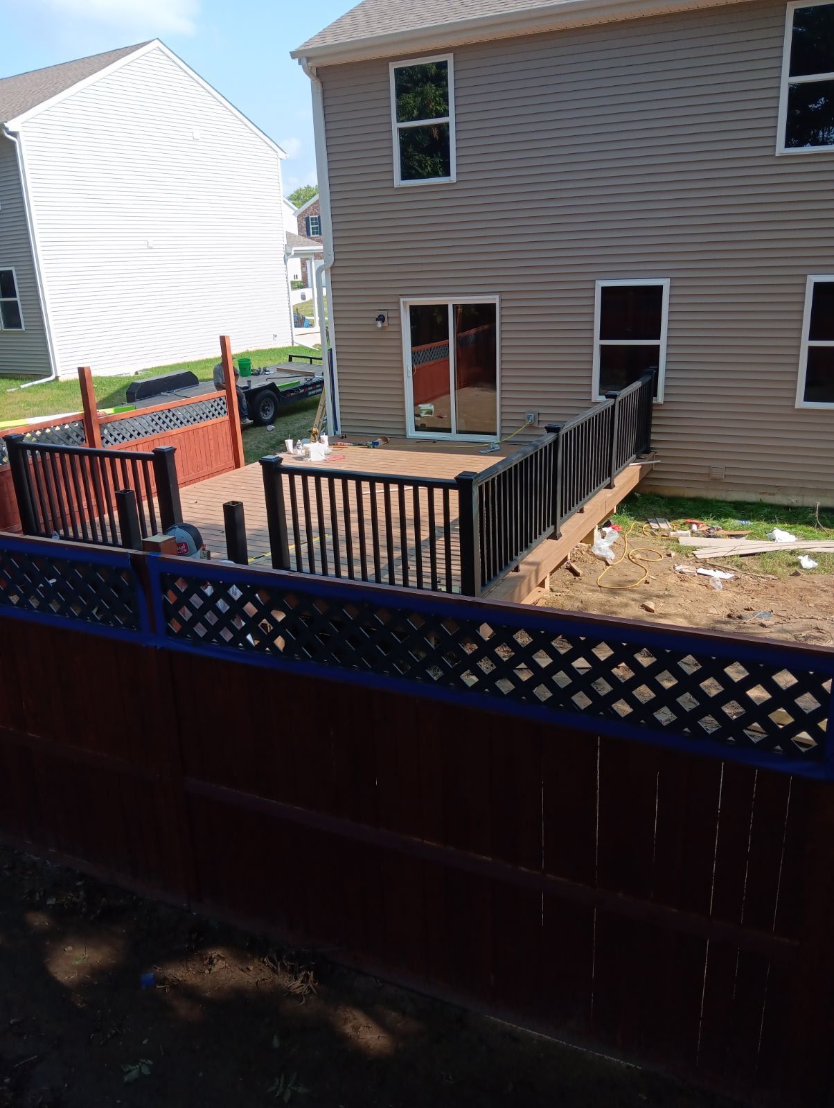 Backyard with a deck, fence, and house. Brown and black wooden features, green grass, clear sky.