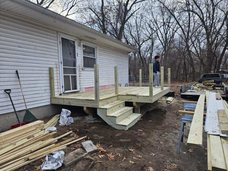 Newly built wooden deck with steps attached to a white house. A person stands on the deck. Construction materials are visible.