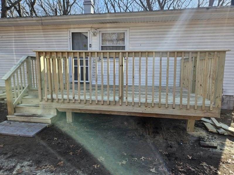 Wooden deck with railings and steps attached to a white building. Ground is dirt and some debris.