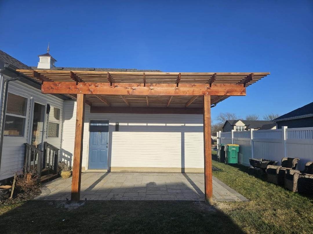 Wooden pergola attached to a white building; blue door below. Sunny outdoors.