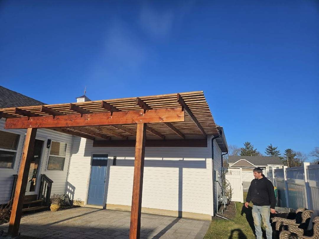 Wooden carport next to a white building; a person stands nearby on a sunny day.
