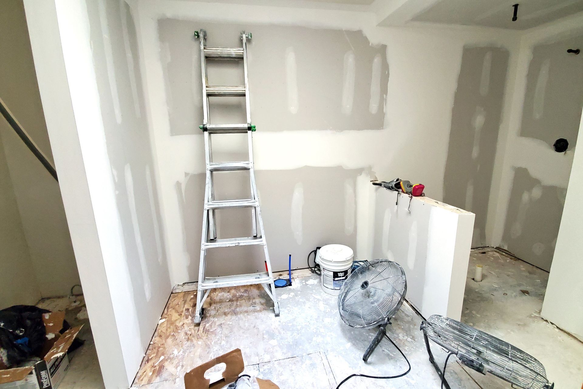 Interior room under construction, drywall installed. Ladder, fan, and supplies present.