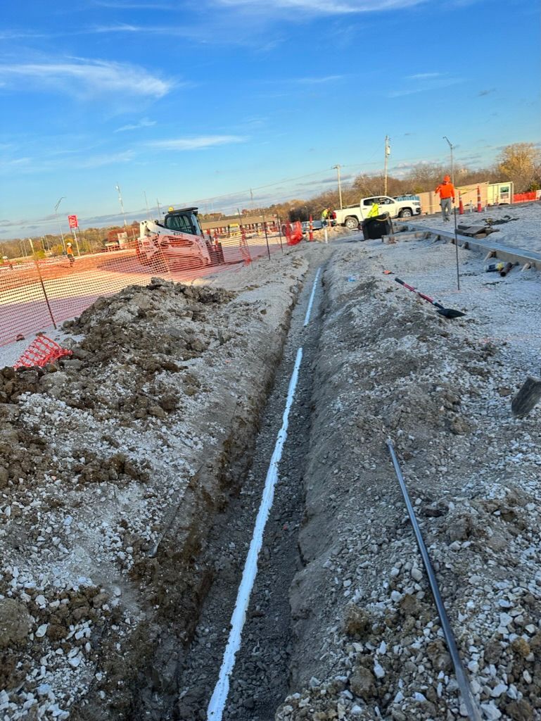 Cowboys Plumbing - A trench with a white pipe, construction site with workers and vehicles under a blue sky.