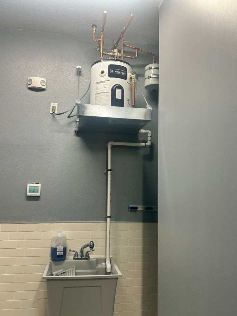Cowboys Plumbing - Water heater, pipes, and sink in a utility room. Gray and white, with a bottle of soap on the sink.