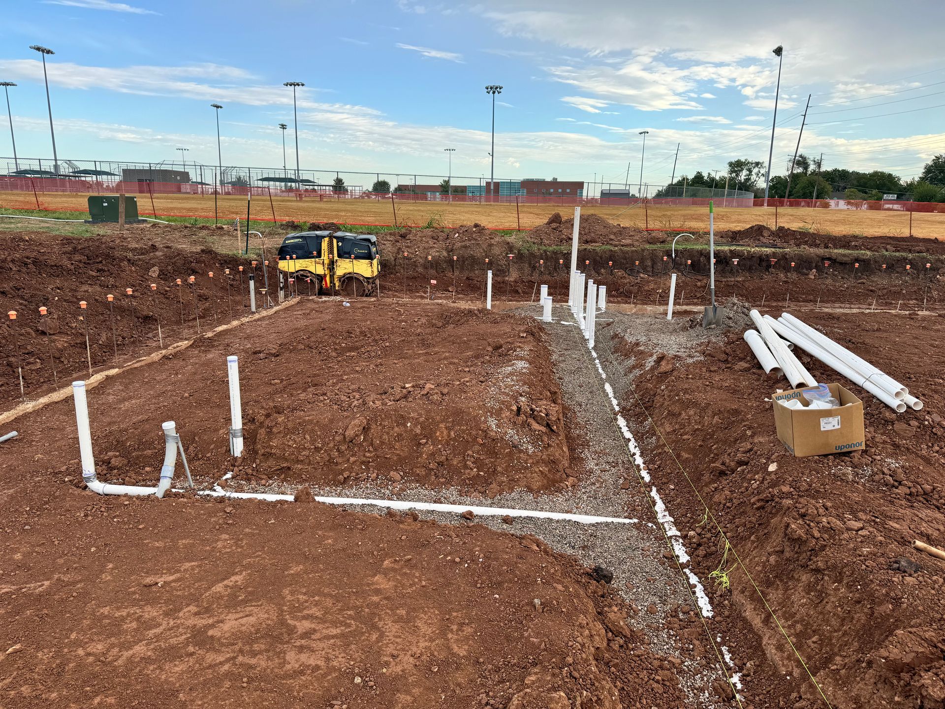 Cowboys Plumbing - Construction site: trench with PVC pipes and gravel, bulldozer, red soil, blue sky.
