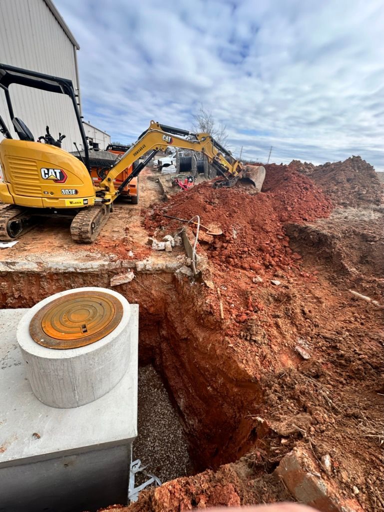 Cowboys Plumbing - Construction site: excavator digging red dirt near concrete structures and a building under a cloudy sky.