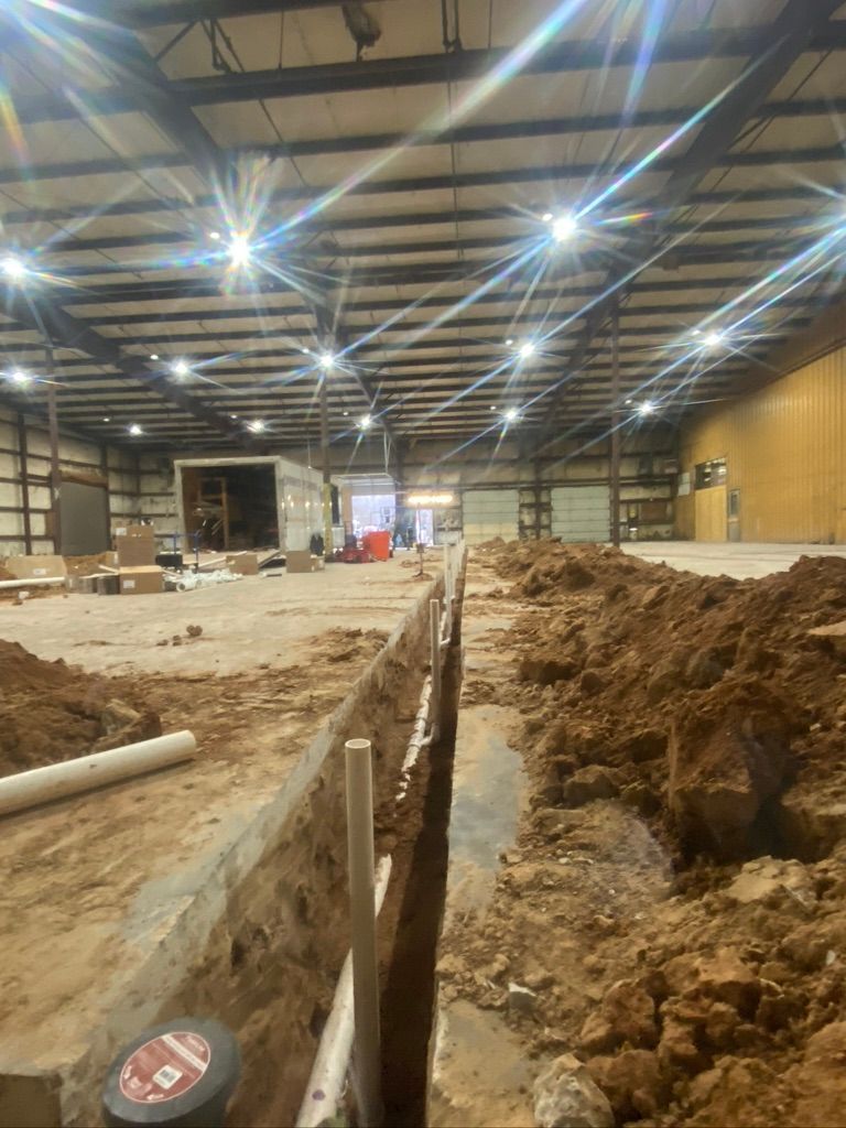 Cowboys Plumbing - Construction site inside a warehouse. A trench is dug with PVC pipes visible; dirt piles surround the trench.