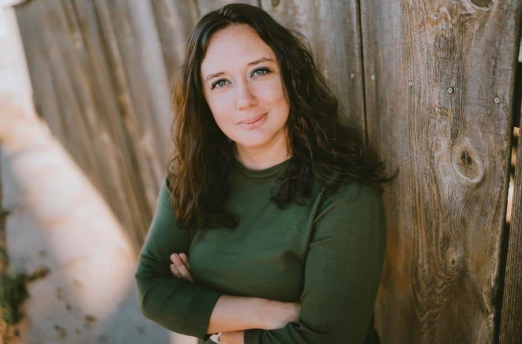 Woman with crossed arms, wearing green shirt, leans against a wooden fence; smiling, outdoors.
