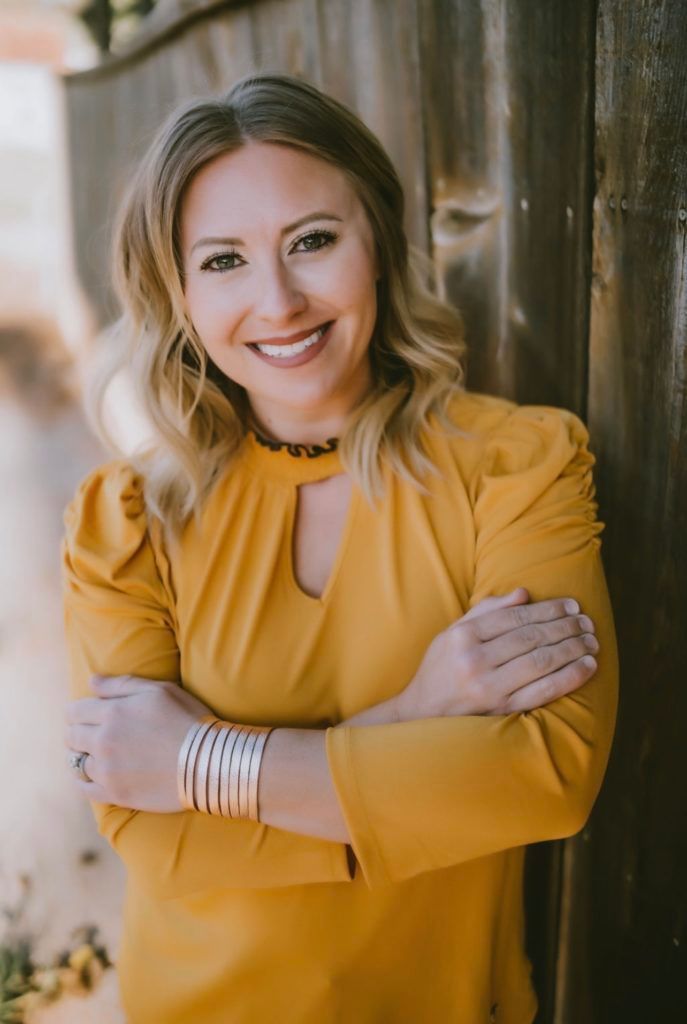 Woman with blonde hair smiles, arms crossed, wearing a yellow blouse, leaning against a wooden fence.