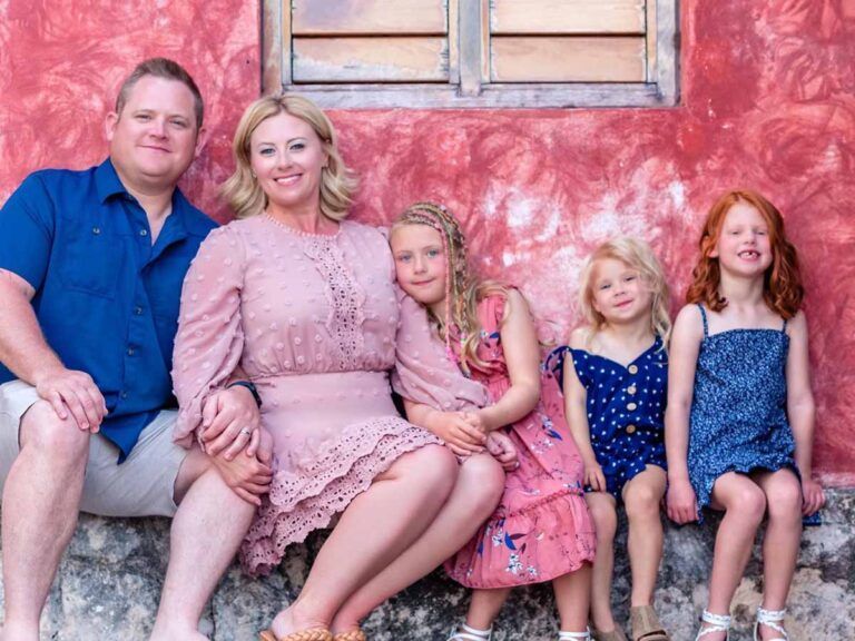 Family of five, seated against a red wall; parents smiling, three daughters with different hair colors.