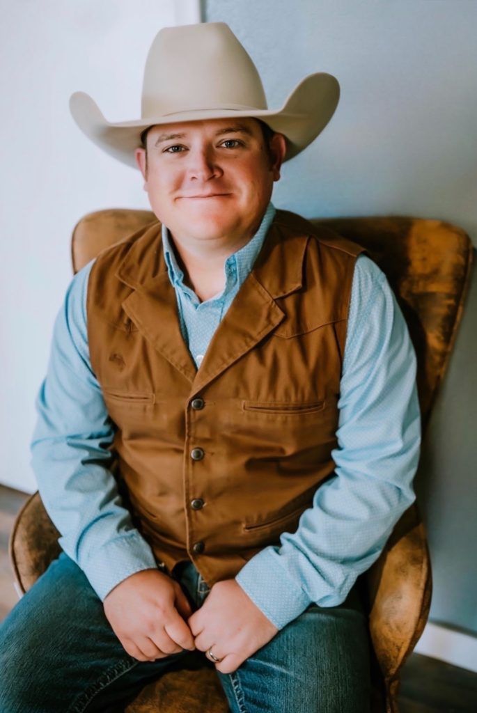 Man in cowboy hat and vest seated in a chair, smiling.