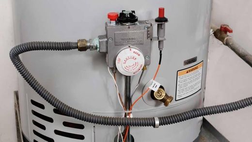 Close-up of a gas water heater control panel, showing the thermostat dial, pilot light button, and gas line connection.