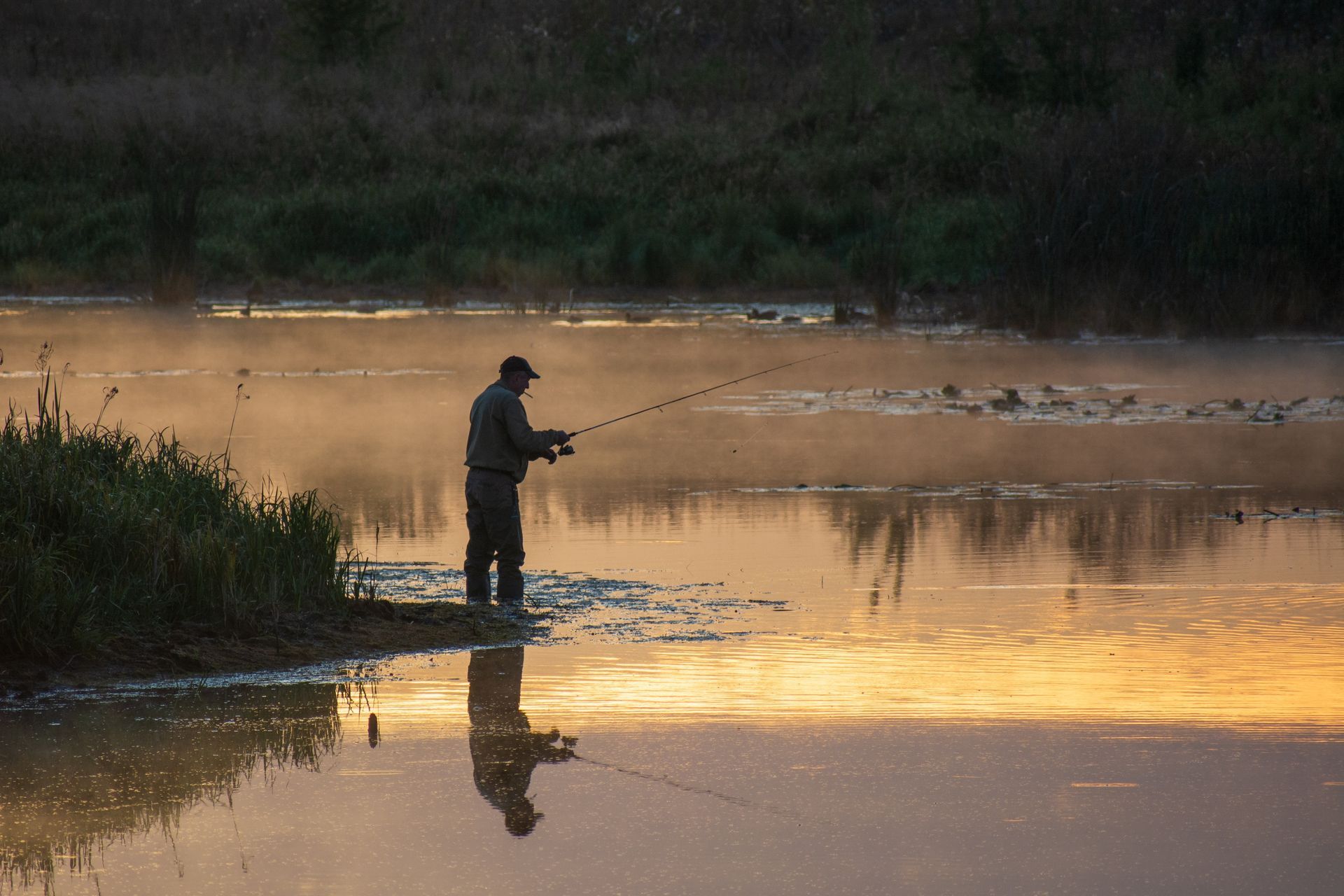 A lone person stands at the water's edge at sunrise, fishing with a pole while mist drifts over the calm, golden lake.