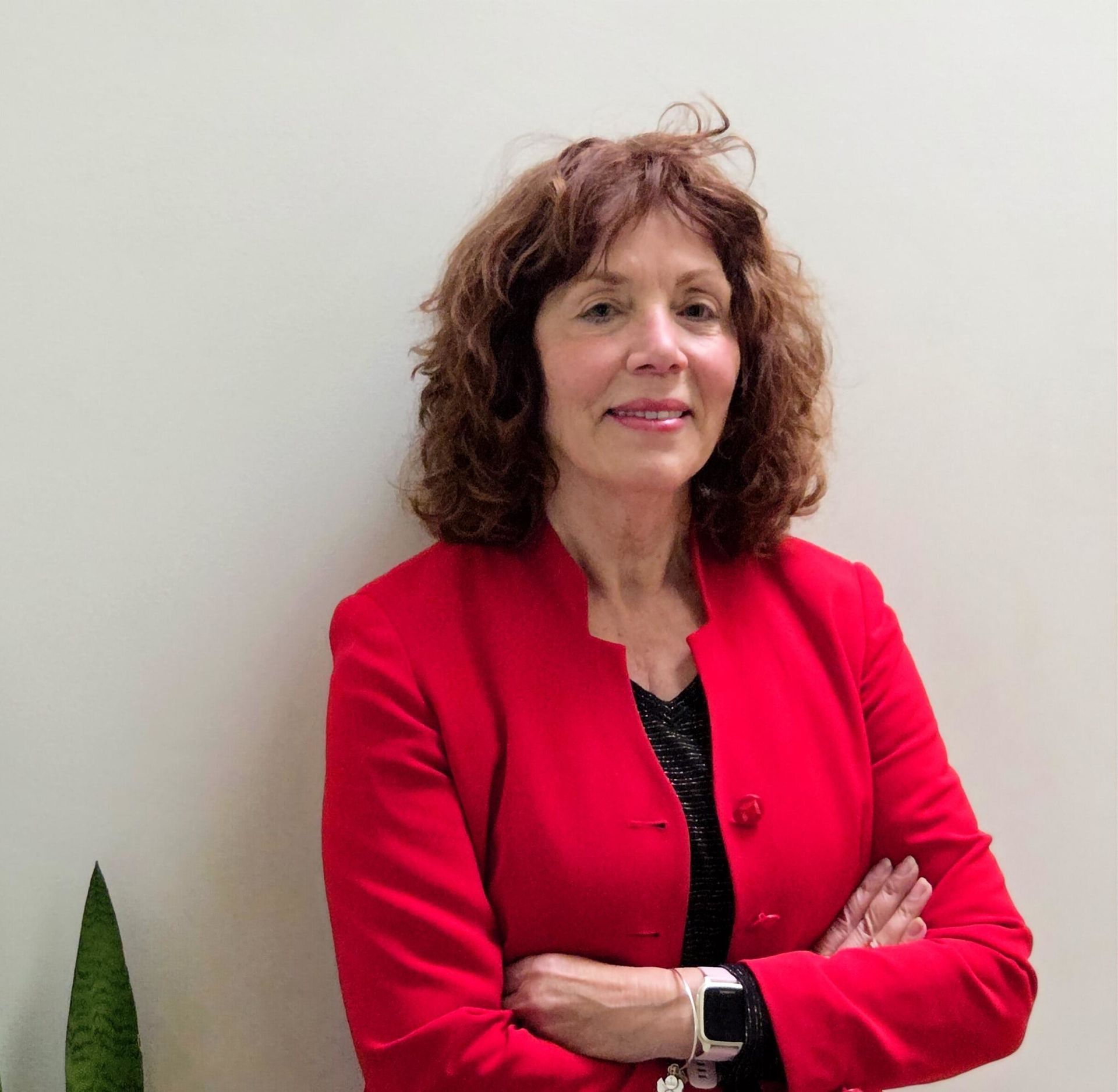 Woman in a red blazer standing with arms crossed against a plain light wall