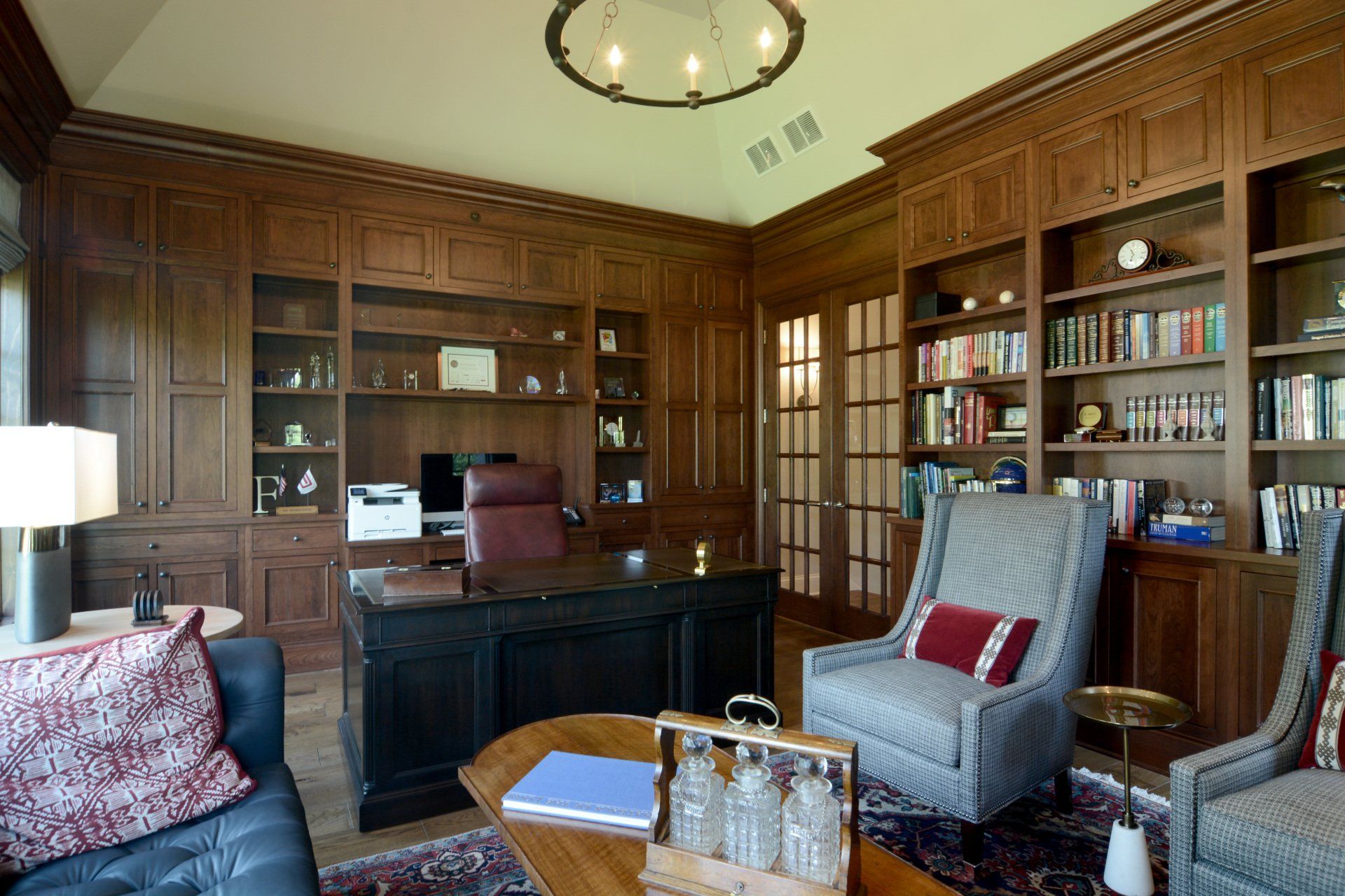 A living room with a desk , chairs , bookshelves and a chandelier.