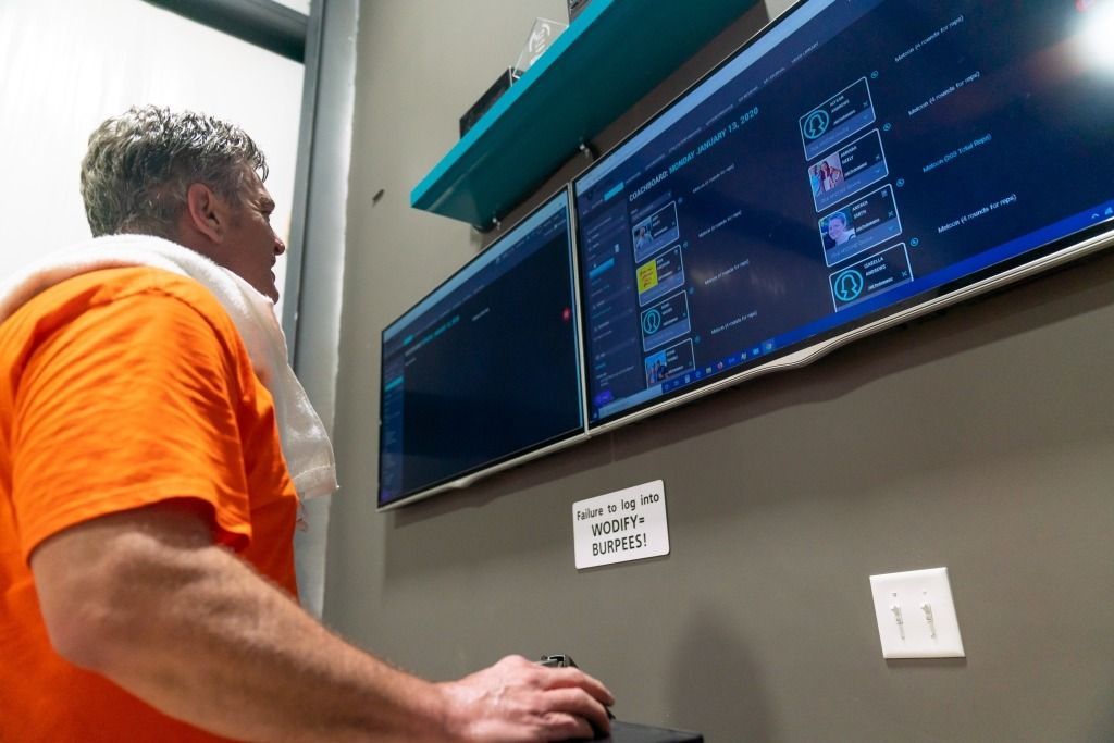 A man in an orange shirt is standing in front of a computer monitor.