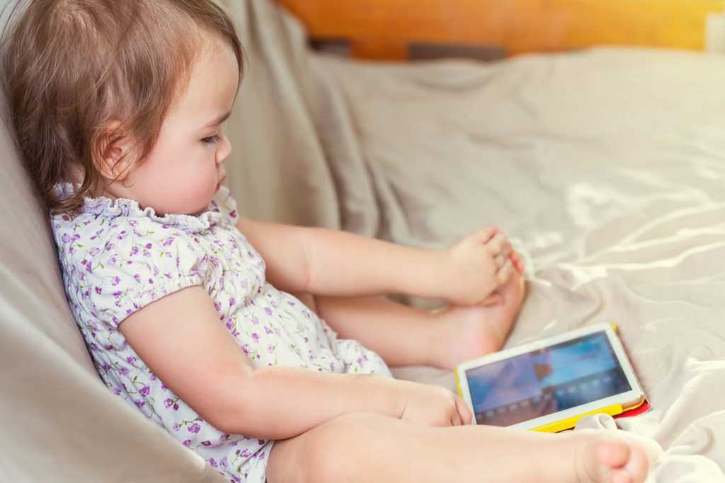 A little girl is sitting on a bed playing with a tablet.