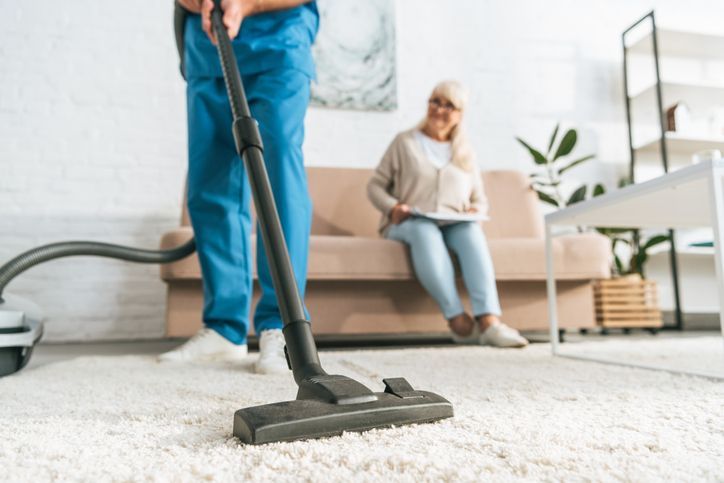 A man is vacuuming the floor while a woman sits on a couch.
