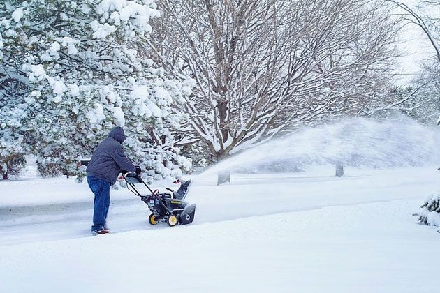 A man is blowing snow from a snow blower on a snowy driveway.