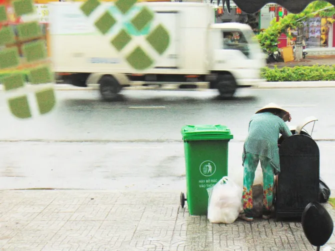 Person standing by a green bin as a garbage truck passes by on a city street.