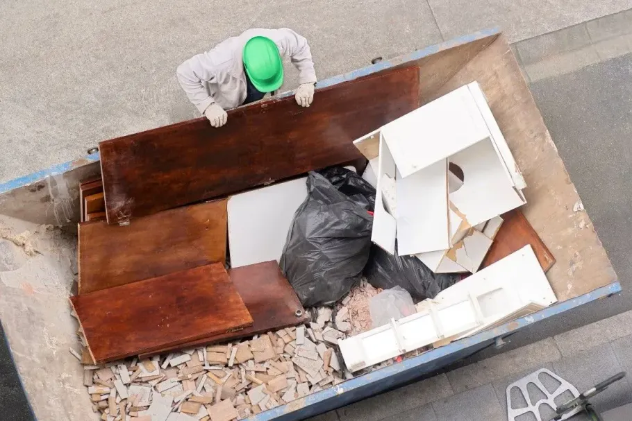 Worker in green helmet loads wood and trash into a large dumpster.
