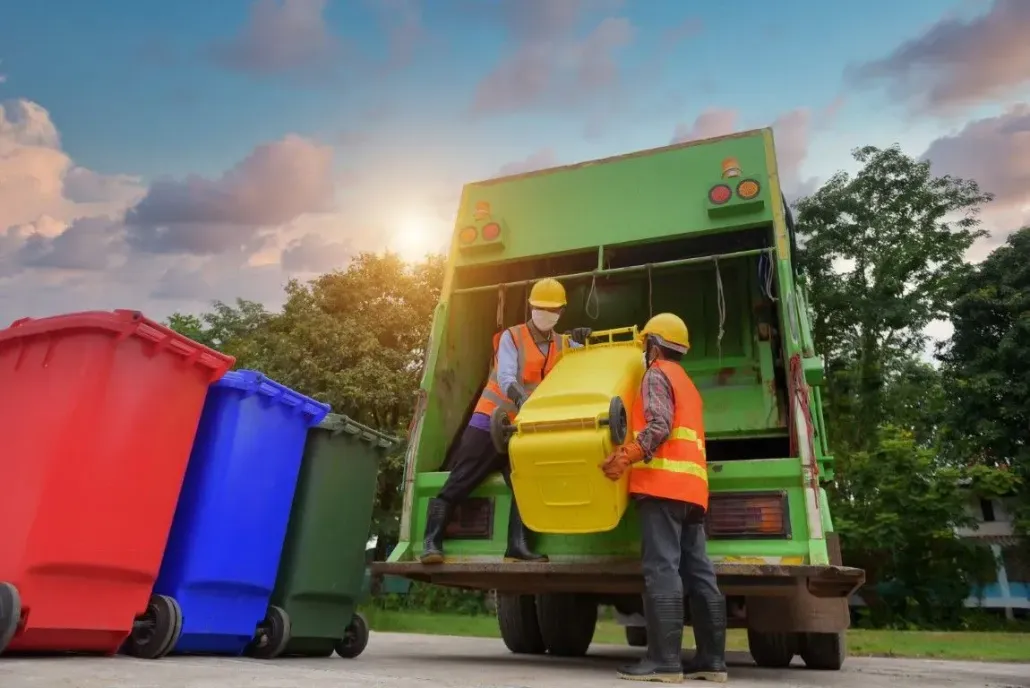 Workers in safety gear emptying large trash bins into a green garbage truck.
