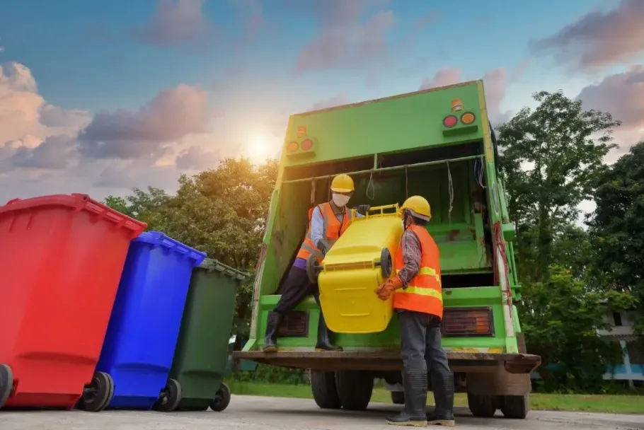 Garbage workers emptying a yellow bin into a green truck beside blue and red bins at sunset