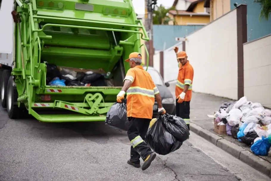 Garbage collectors in orange uniforms loading black trash bags into a green truck on a street